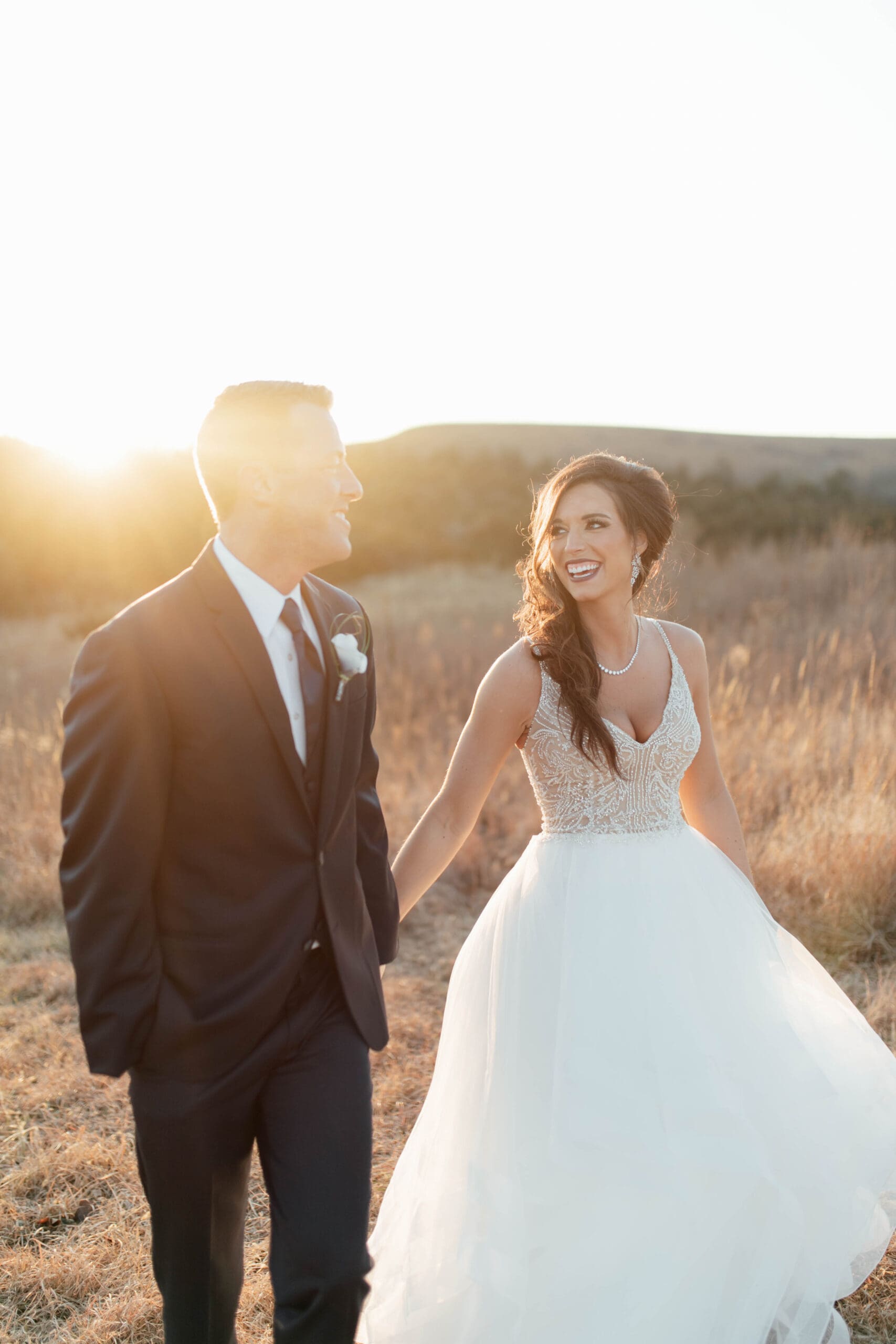 Bride and groom walking hand-in-hand through a golden field at sunset, smiling joyfully on their wedding day.