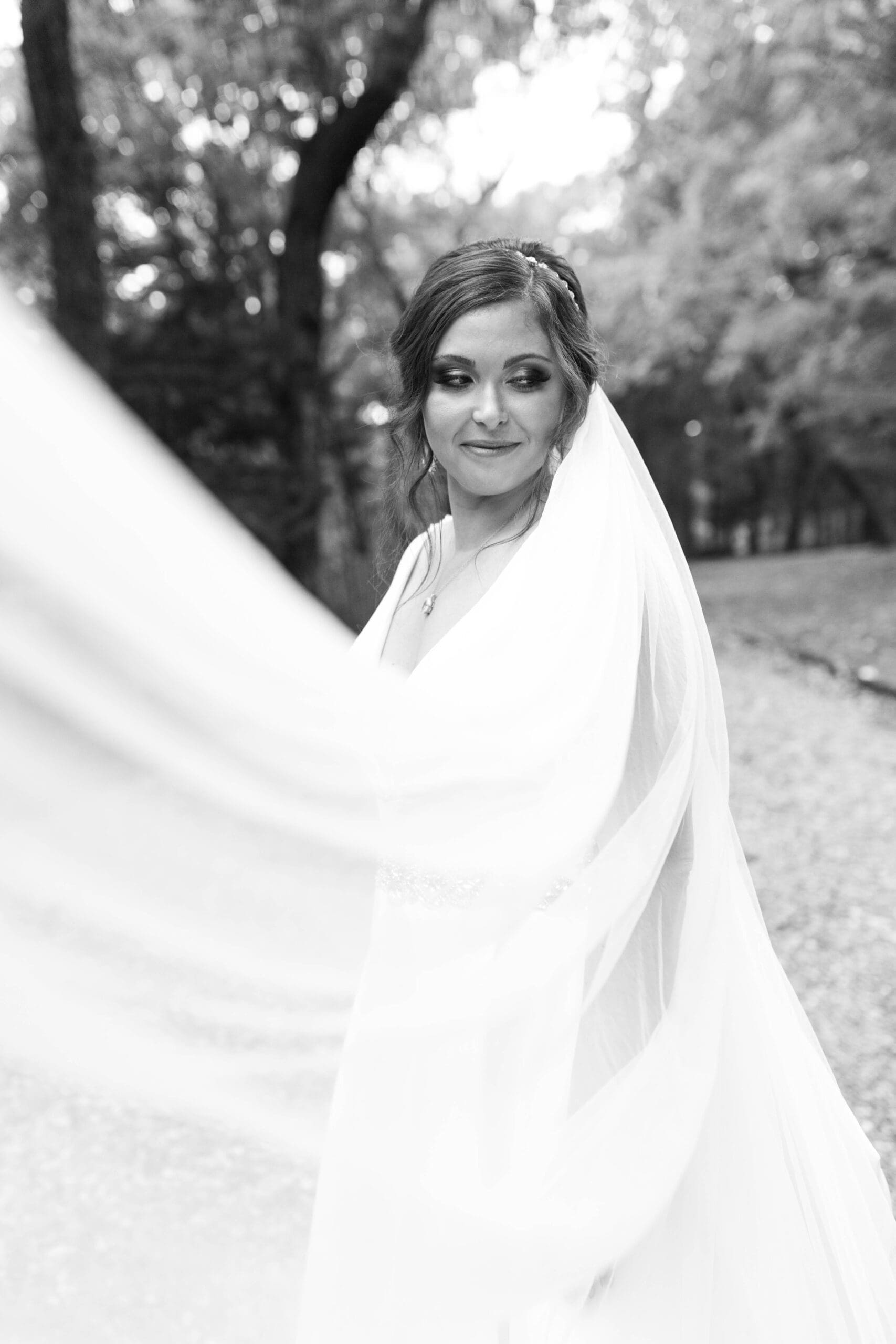 Black and white photo of a bride smiling softly, with her veil flowing around her in a wooded outdoor setting.