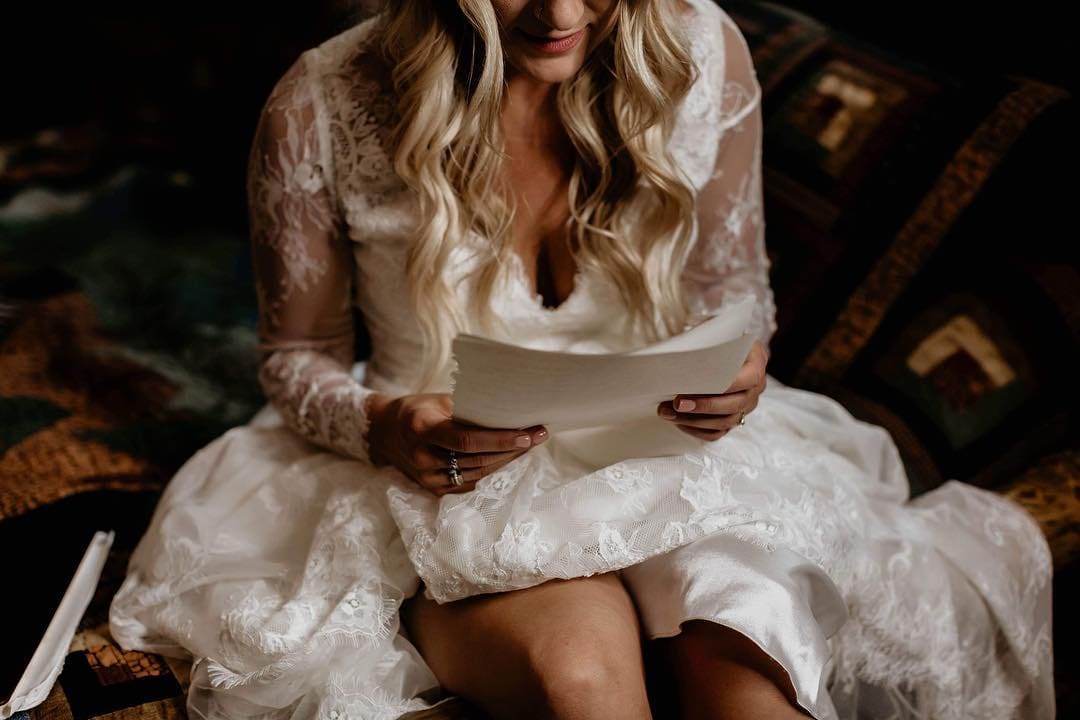 A bride in a lace wedding dress sits and reads a handwritten letter, smiling softly, with natural light highlighting her wavy blonde hair.