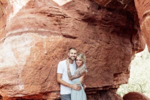 Engaged couple standing in front of red rock cliffs at Garden of the Gods, holding hands.