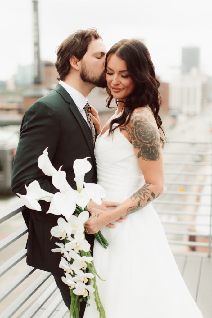 Bride and groom on rooftop while groom kisses bride's cheek.