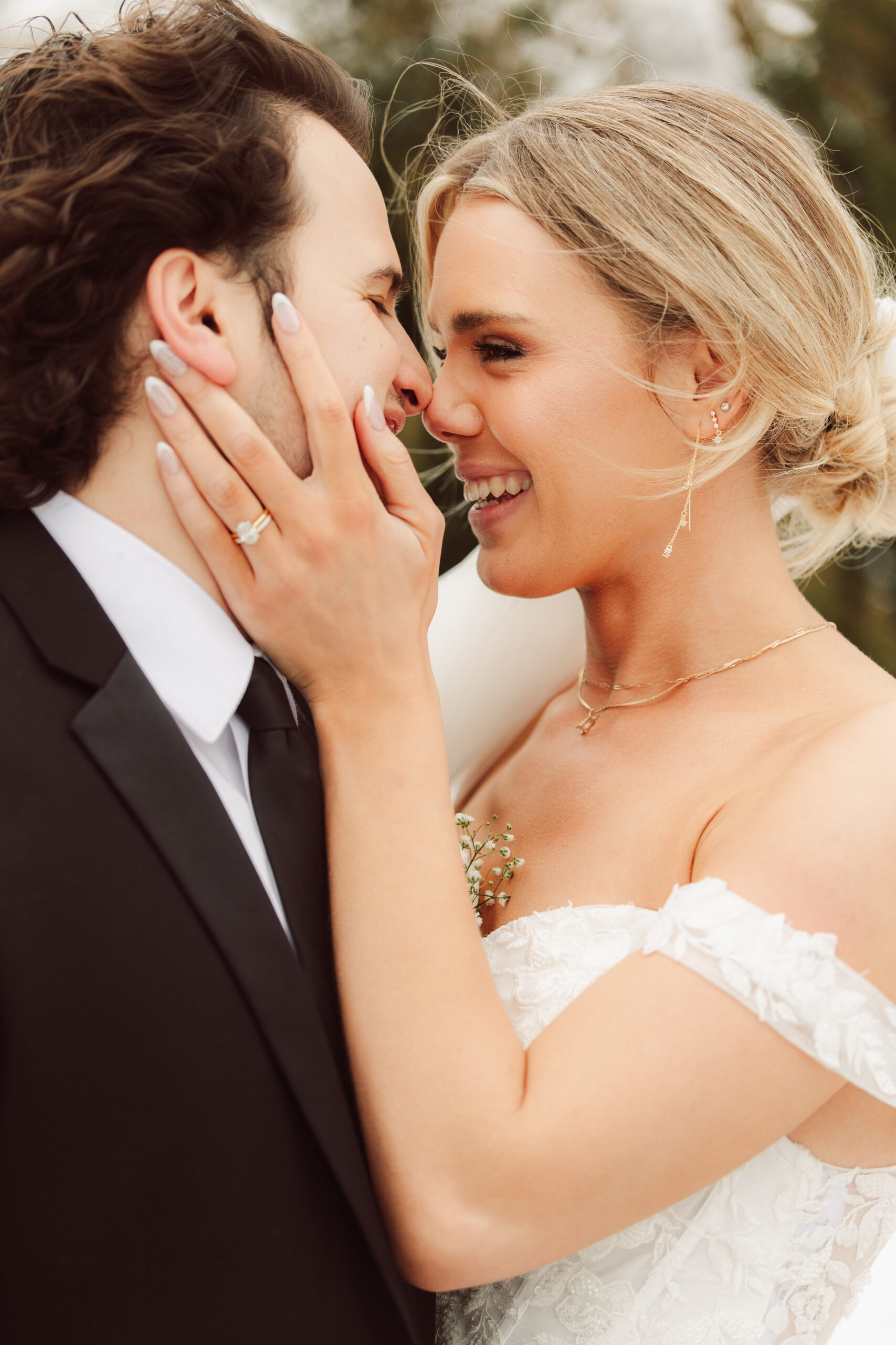 Bride gently holding groom’s face and smiling during an intimate wedding portrait, showcasing her engagement ring and off-the-shoulder dress.