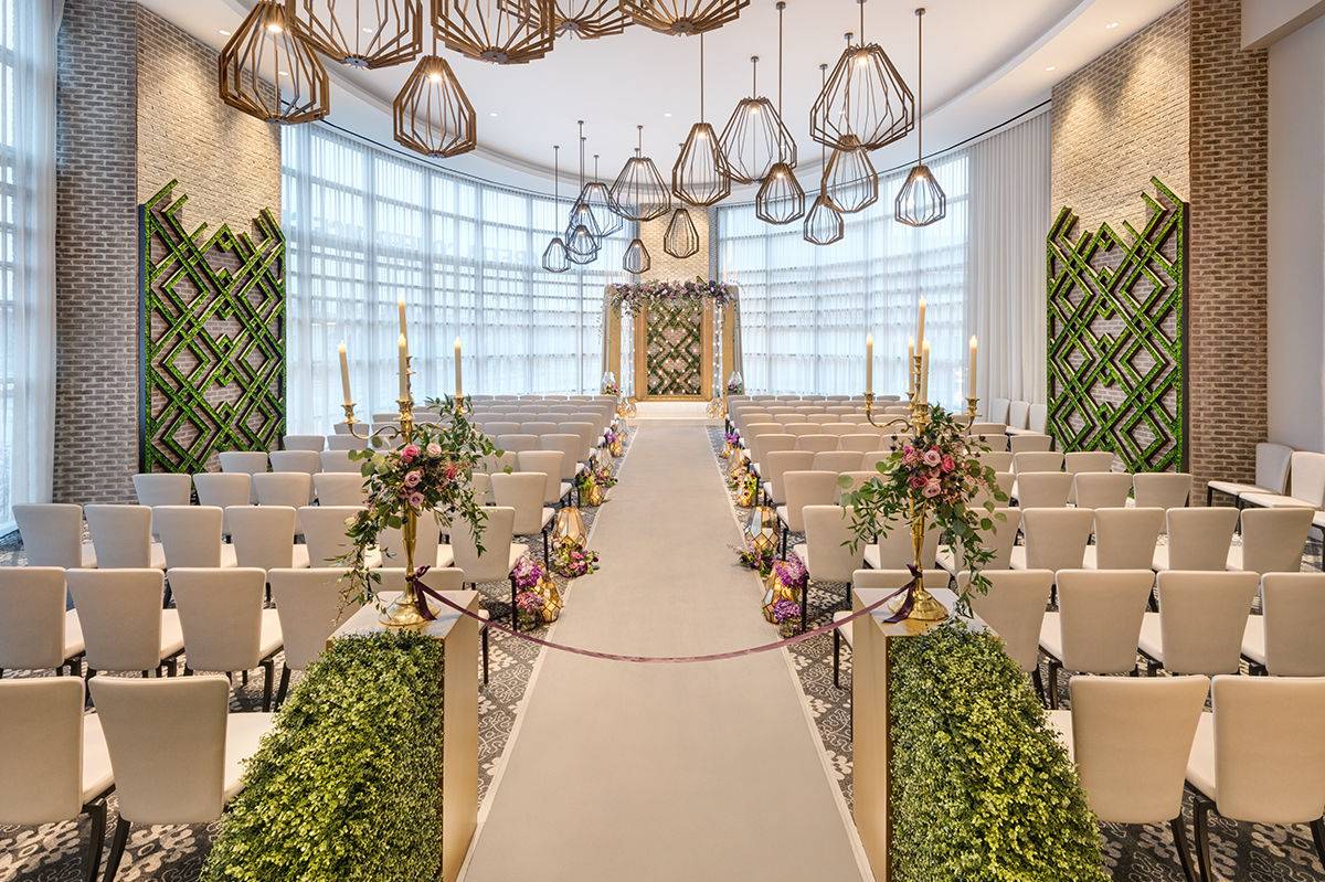 Indoor wedding ceremony space at The Hotel at Avalon in Alpharetta, Georgia featuring modern chandeliers, floor-to-ceiling windows, floral aisle arrangements, and elegant neutral seating.